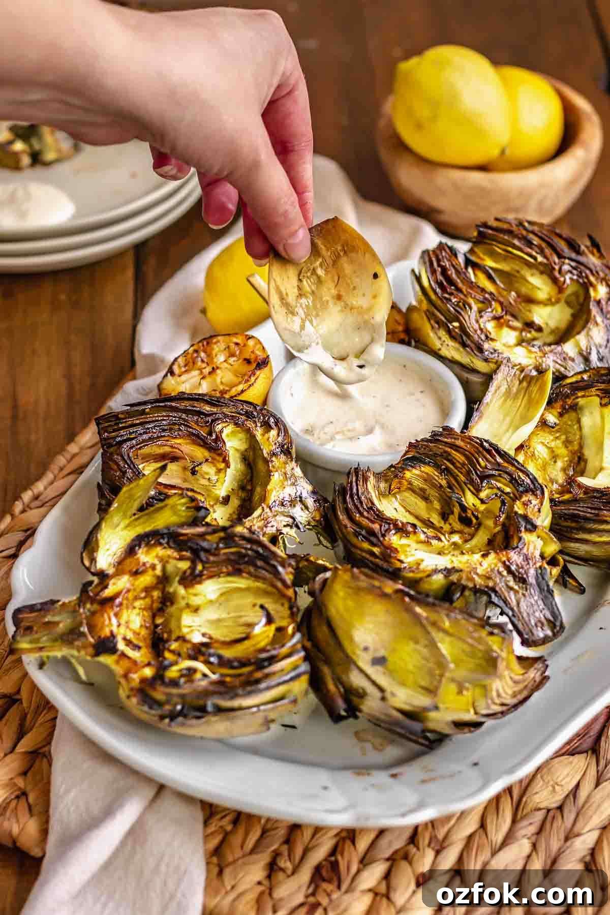 Grilled Marinated Artichokes with Lemon Aioli Recipe 2 A hand dips an artichoke leaf into aioli. Other artichokes sit on a platter.