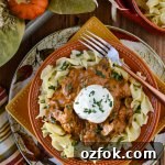Beef Stroganoff on a bed of buttered egg noodles in fall dinnerware next to a glass of wine. There is a side dish of noodles and striped napkin.