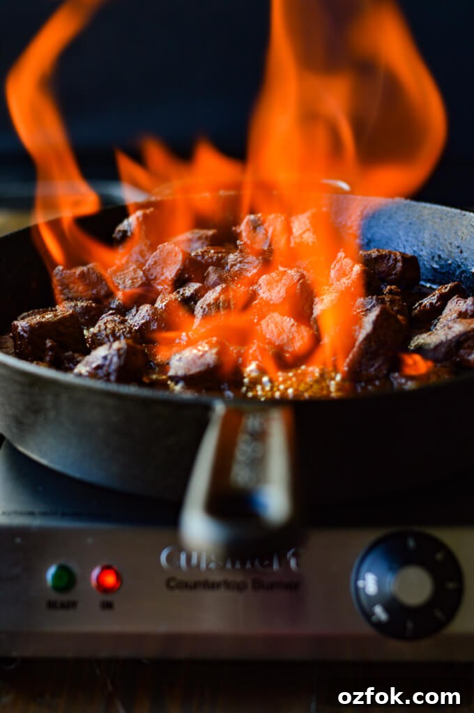 A front view of a cast iron skillet with flambé beef stroganoff while the fire is flaring.