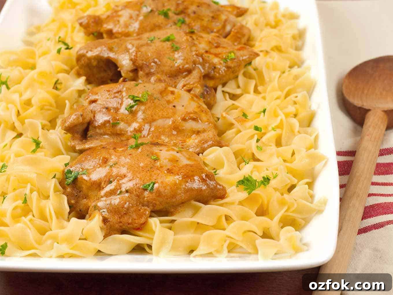 Classic Hungarian Chicken Paprikash Recipe — Authentic Paprikas 2 Easy Chicken Paprikas served over a bed of buttered egg noodles, garnished with chopped parsley on a rectangular white platter.