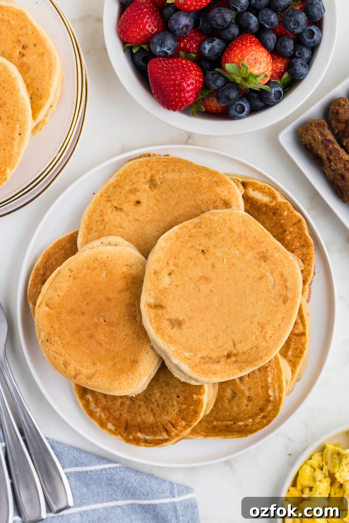 Light and fluffy pancakes stacked on a plate with fruit and sausage on the table. 