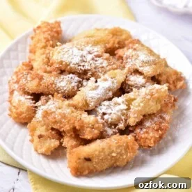A plate of crispy banana tempura dusted with powdered sugar sits elegantly on a white plate atop a yellow cloth.