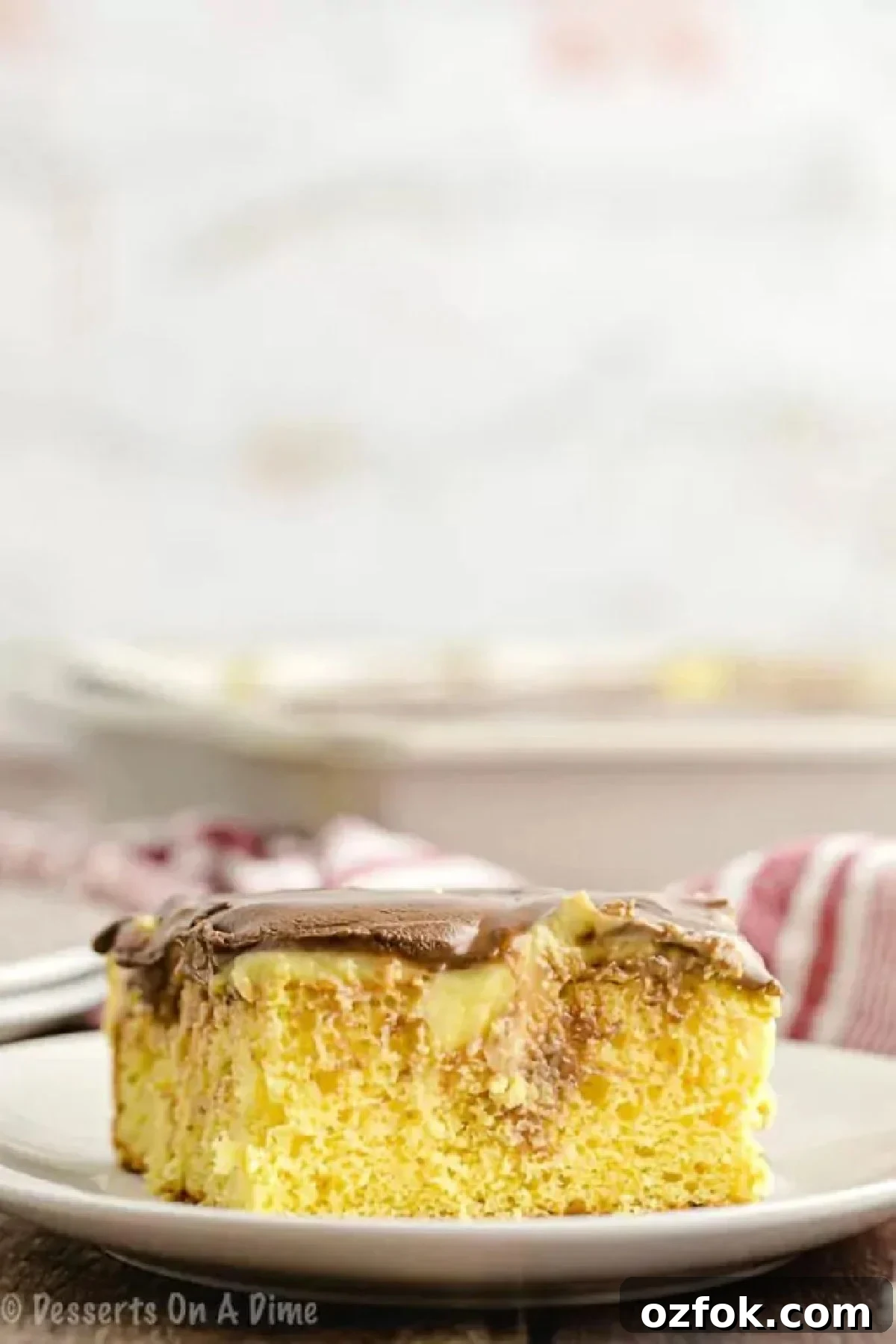 Boston creme poke cake recipe is so decadent and full of flavor. Make this easy cake in minutes with only a few ingredients for a delicious dessert. A slice of the Boston Cream Poke Cake on a white serving plate.
