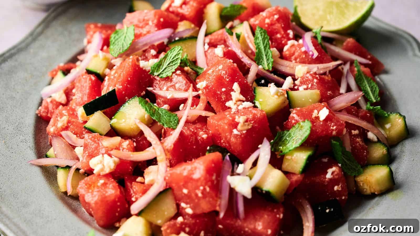 29 Irresistible Salad Recipes Everyone Will Love 26 A plate of watermelon salad with cucumber, red onion, feta cheese, and mint leaves, served with a lime wedge.