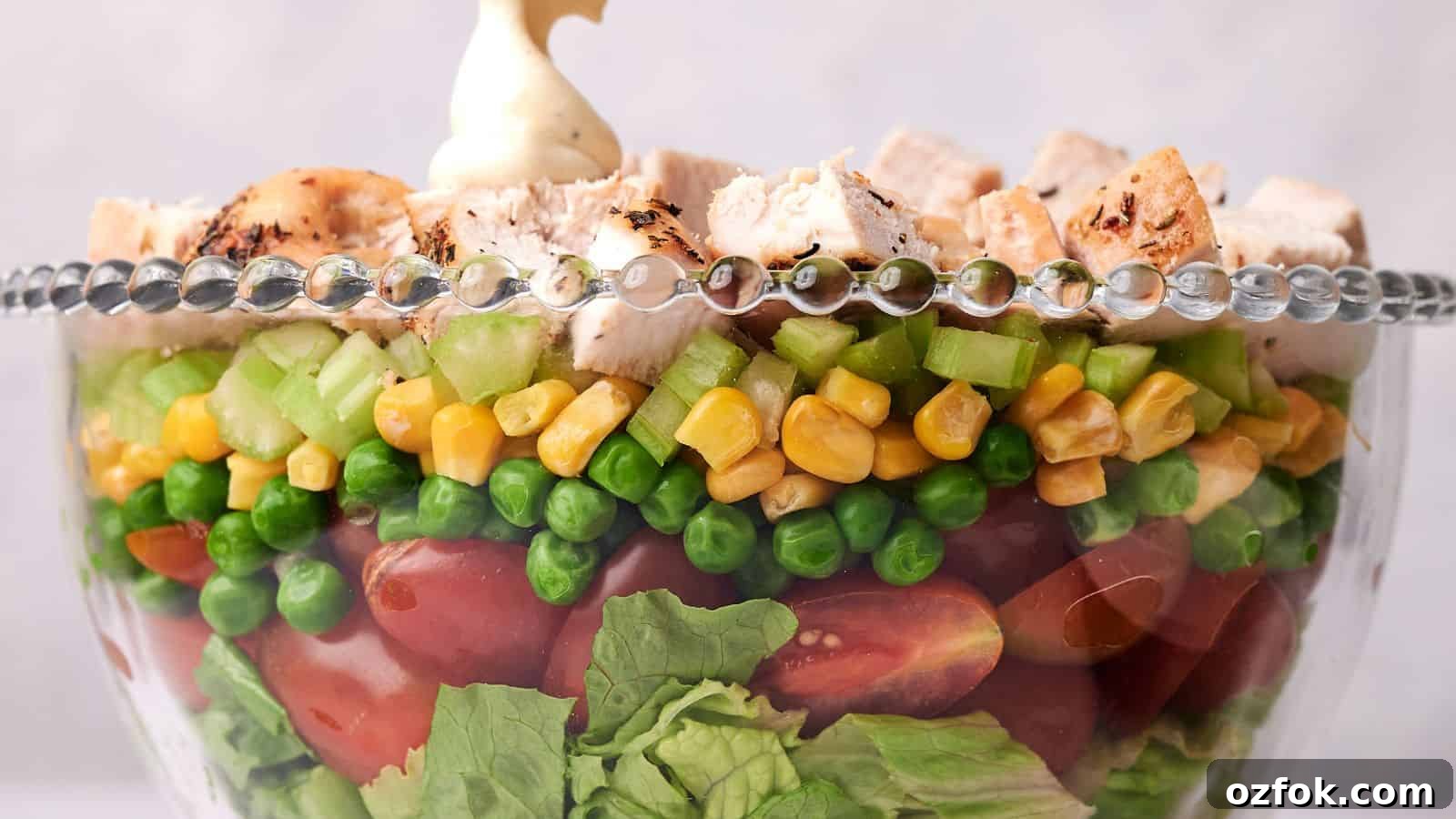 29 Irresistible Salad Recipes Everyone Will Love 10 A clear glass bowl layered with lettuce, cherry tomatoes, green peas, corn, diced celery, and topped with chunks of grilled chicken, garnished with a dollop of mayonnaise.