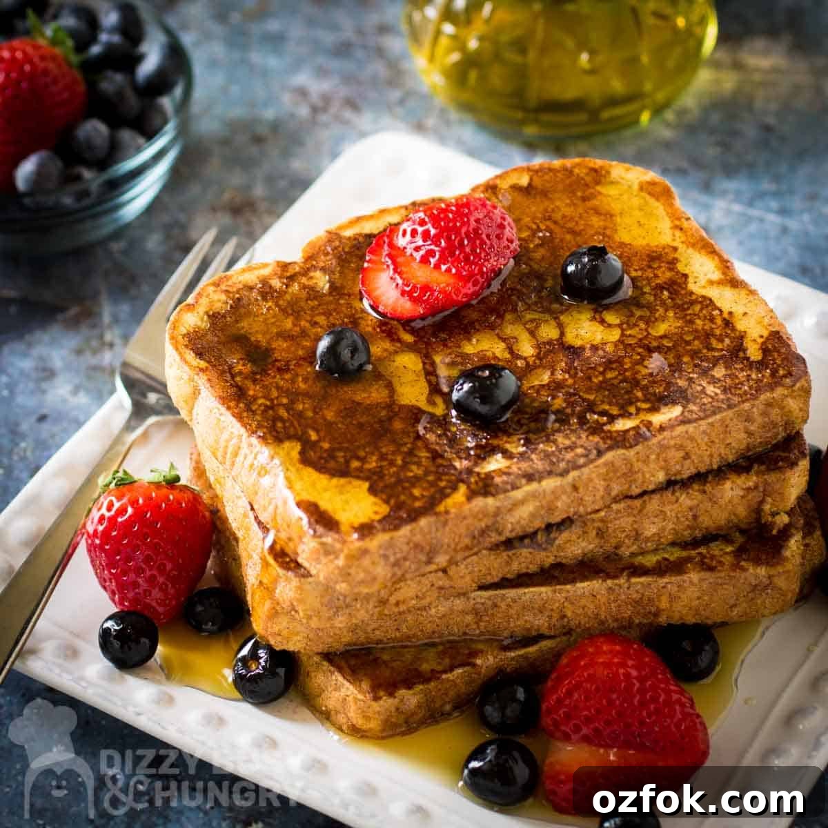 Side shot of french toast stacked on a white plate garnished with sliced strawberries, blueberries, and syrup with a fork on the side.