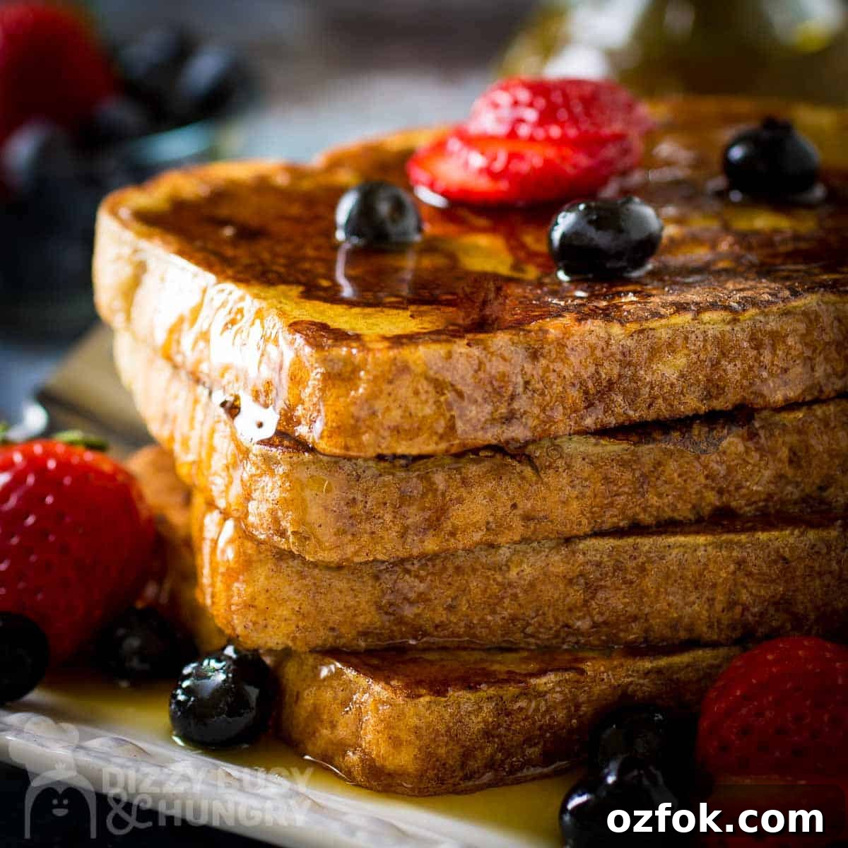 Side close up shot of french toast stacked on a white plate garnished with sliced strawberries, blueberries, and syrup with a fork on the side.