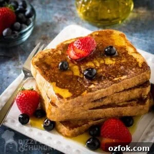 Side shot of french toast stacked on a white plate garnished with sliced strawberries, blueberries, and syrup with a fork on the side.