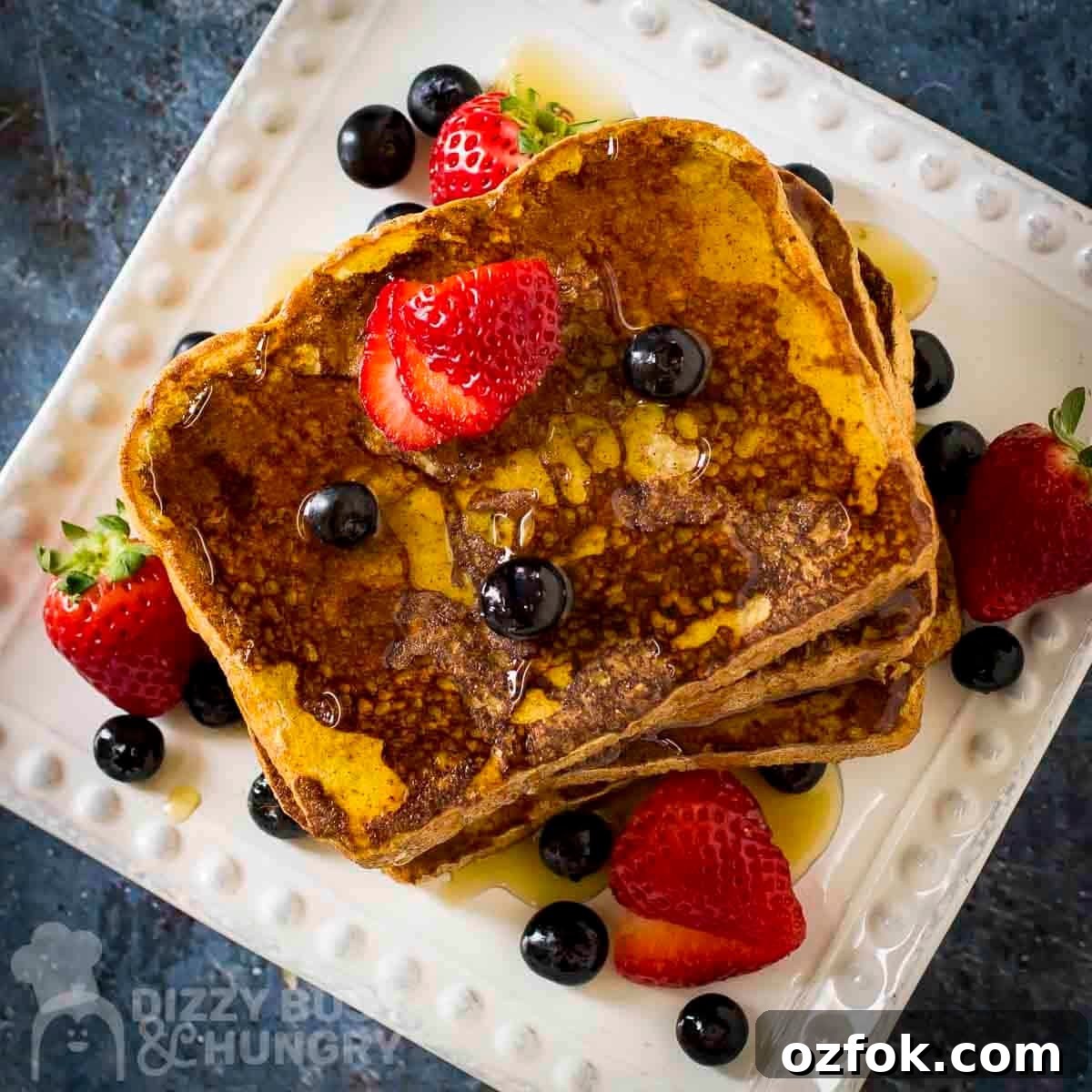 Overhead shot of french toast stacked on a white square plate garnished with sliced strawberries, blueberries, and syrup.