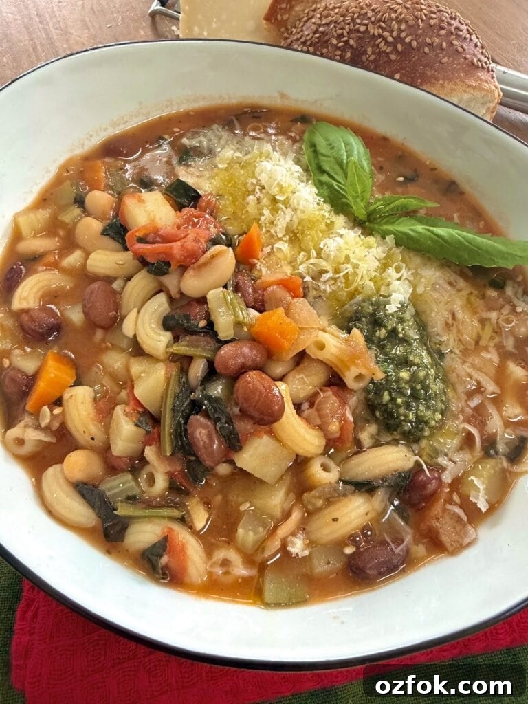 Classic Italian Minestrone Soup Recipe — Hearty Vegetable Stew 2 Italian minestrone soup in white bowl with basil sprig, basil pesto and grated cheese garnish, with seeded bread in the background.