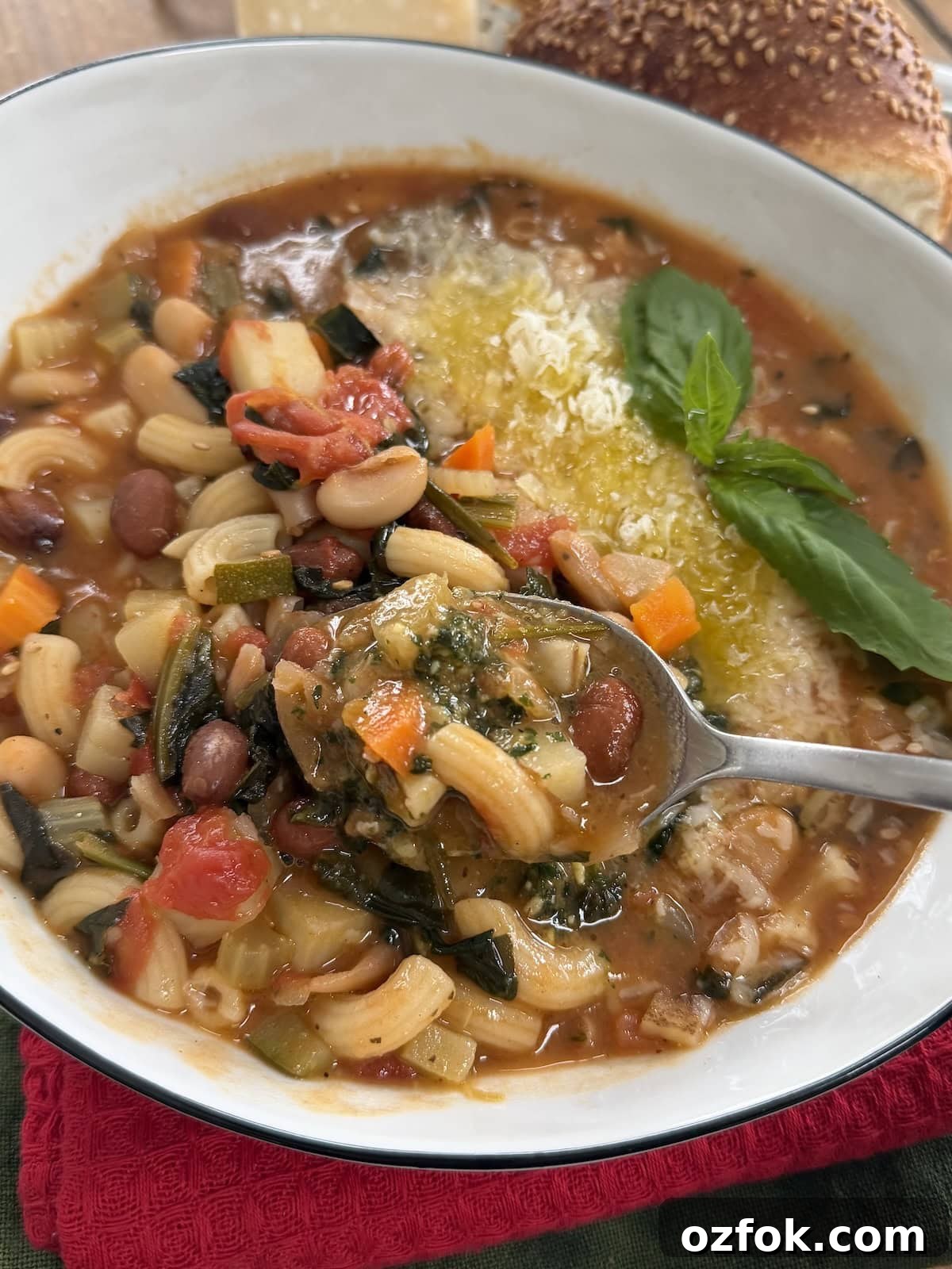 Classic Italian Minestrone Soup Recipe — Hearty Vegetable Stew 13 Italian minestrone soup in white bowl with basil sprig, basil pesto and grated cheese garnish, with seeded bread in the background and a spoon of soup close up.