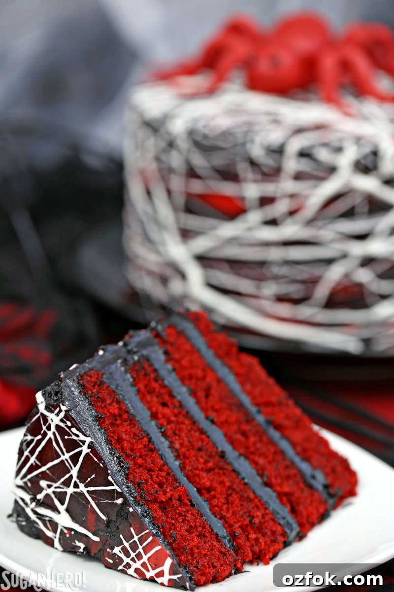 Red Velvet Marshmallow Spiderweb Cake—classic red velvet cake with black chocolate buttercream, covered with a web of marshmallow spiderwebs! Red Velvet Marshmallow Spiderweb Cake - close-up of slice in foreground with whole cake in background