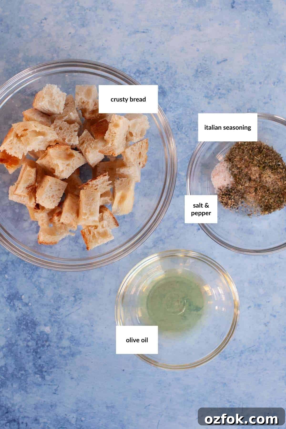 Three bowls contain cubed crusty bread, a mix of Italian seasoning with salt and pepper, and olive oil, all labeled—perfect ingredients for homemade croutons.