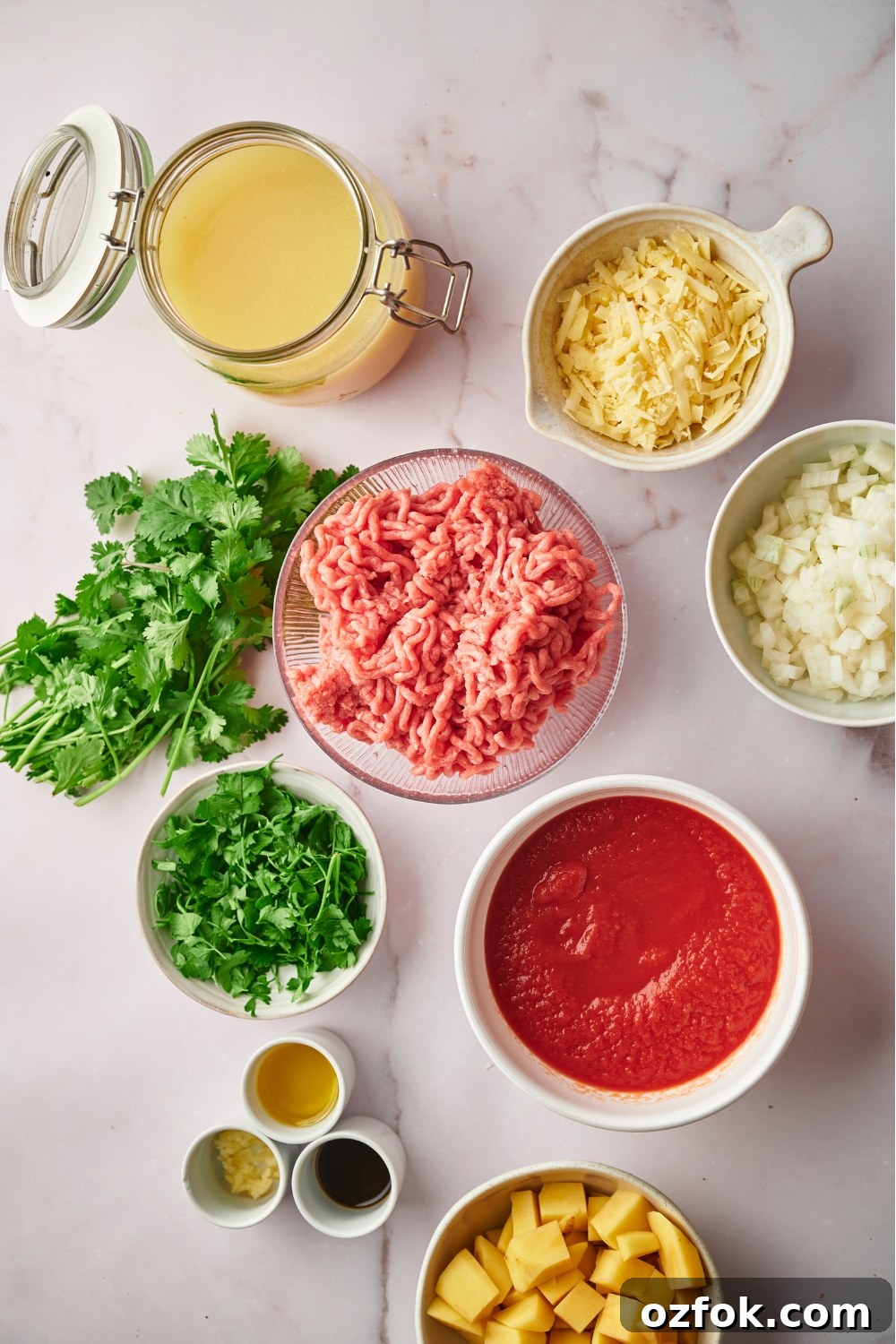 30-Minute Ground Beef and Vegetable Soup Recipe 4 A bowl of ground beef, a glass jar of broth, a bowl of shredded cheese, a bowl of cilantro, a bowl of crushed tomatoes, and part of a bowl of potatoes.