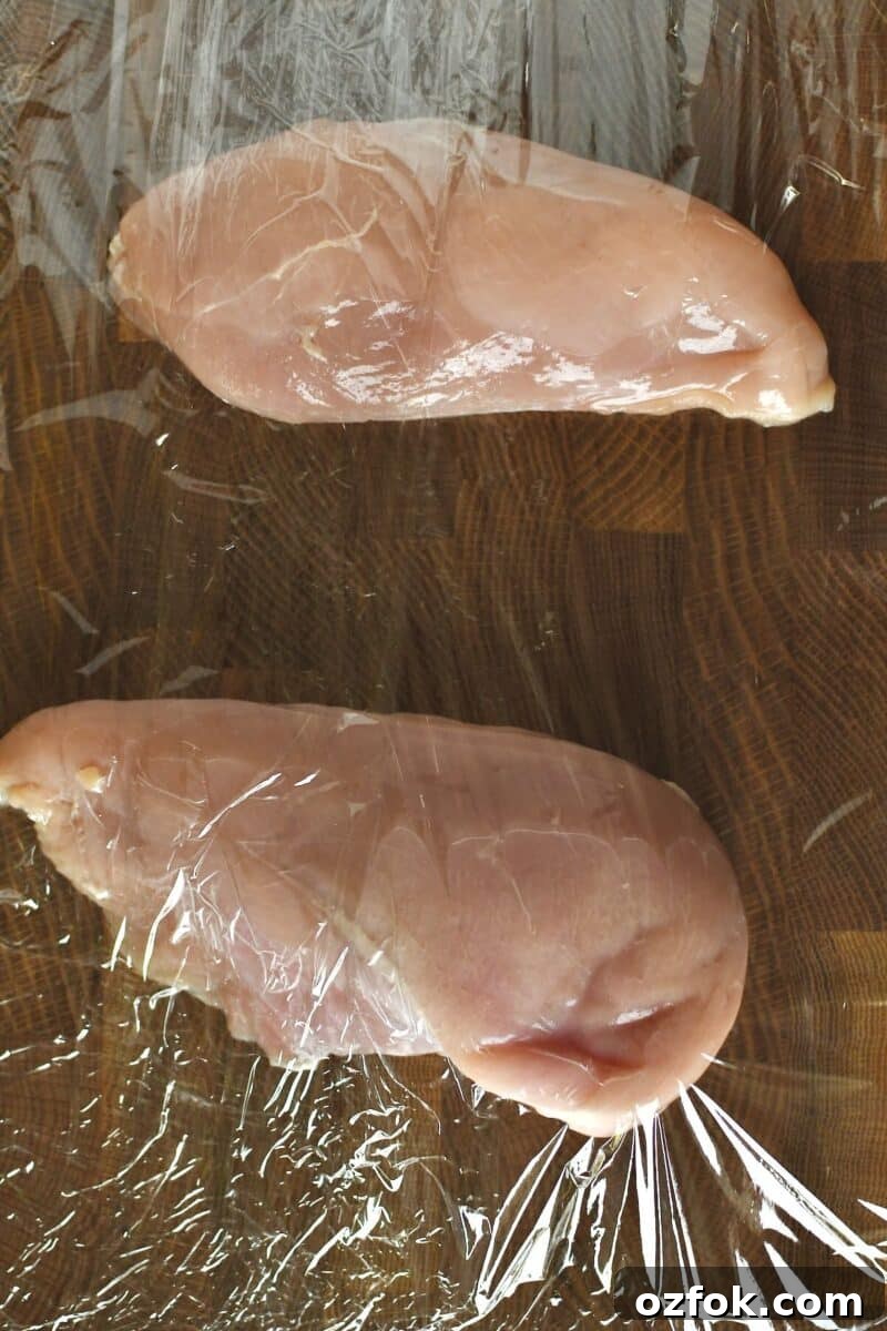 Chicken breasts on a cutting board between two pieces of cling film, before tenderizing to flatten.