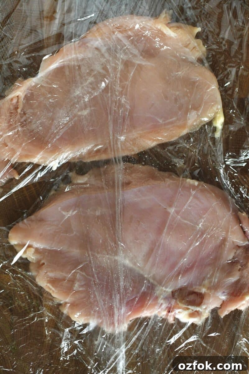 Chicken breasts on a cutting board between two pieces of cling film, after tenderizing to flatten.