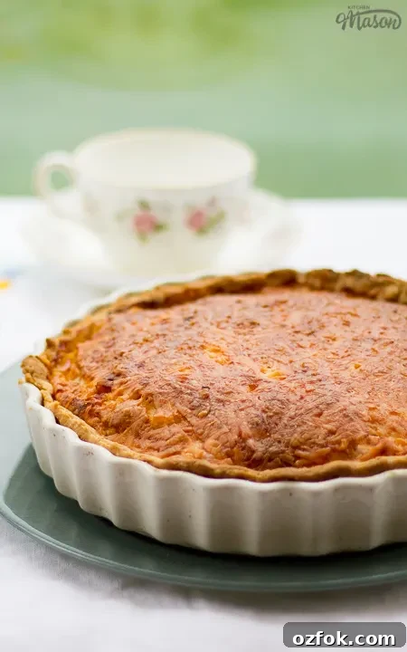Grandma’s Incredible Ham and Cheese Quiche Recipe 2 Gran's ham and cheese quiche in a white baking dish on a green plate. Set on a white floral table cloth with a pastry blender, teacups and a saucer in the background.