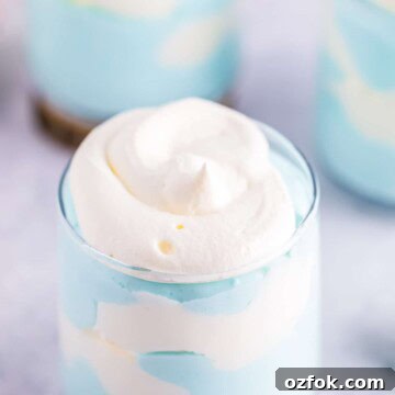 A glass of blue and white jello cloud mousse swirled together like a cloudy sky, topped with whipped cream.