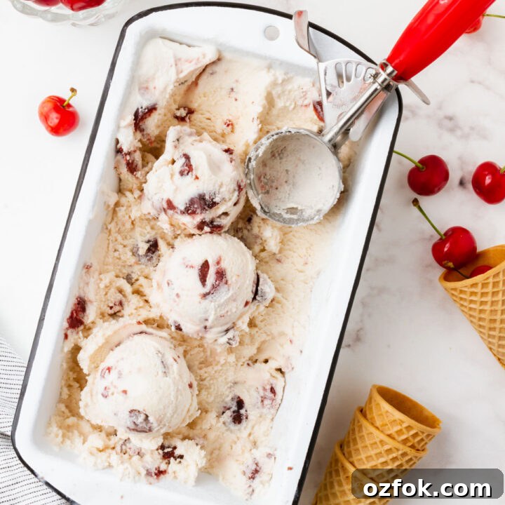 A white loaf pan filled with cherry gelato. A red and metal ice cream scoop is in the pan, and the ice cream has been scooped. Around the pan are fresh cherries and sugar cones.