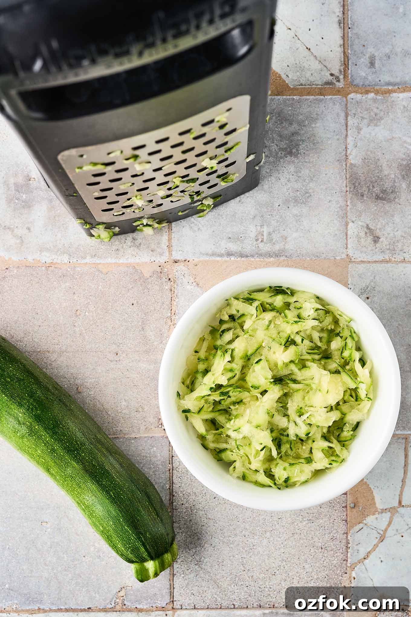 Coarsely grated zucchini in a bowl next to a box grater showing the large holes, a whole zucchini to the side.