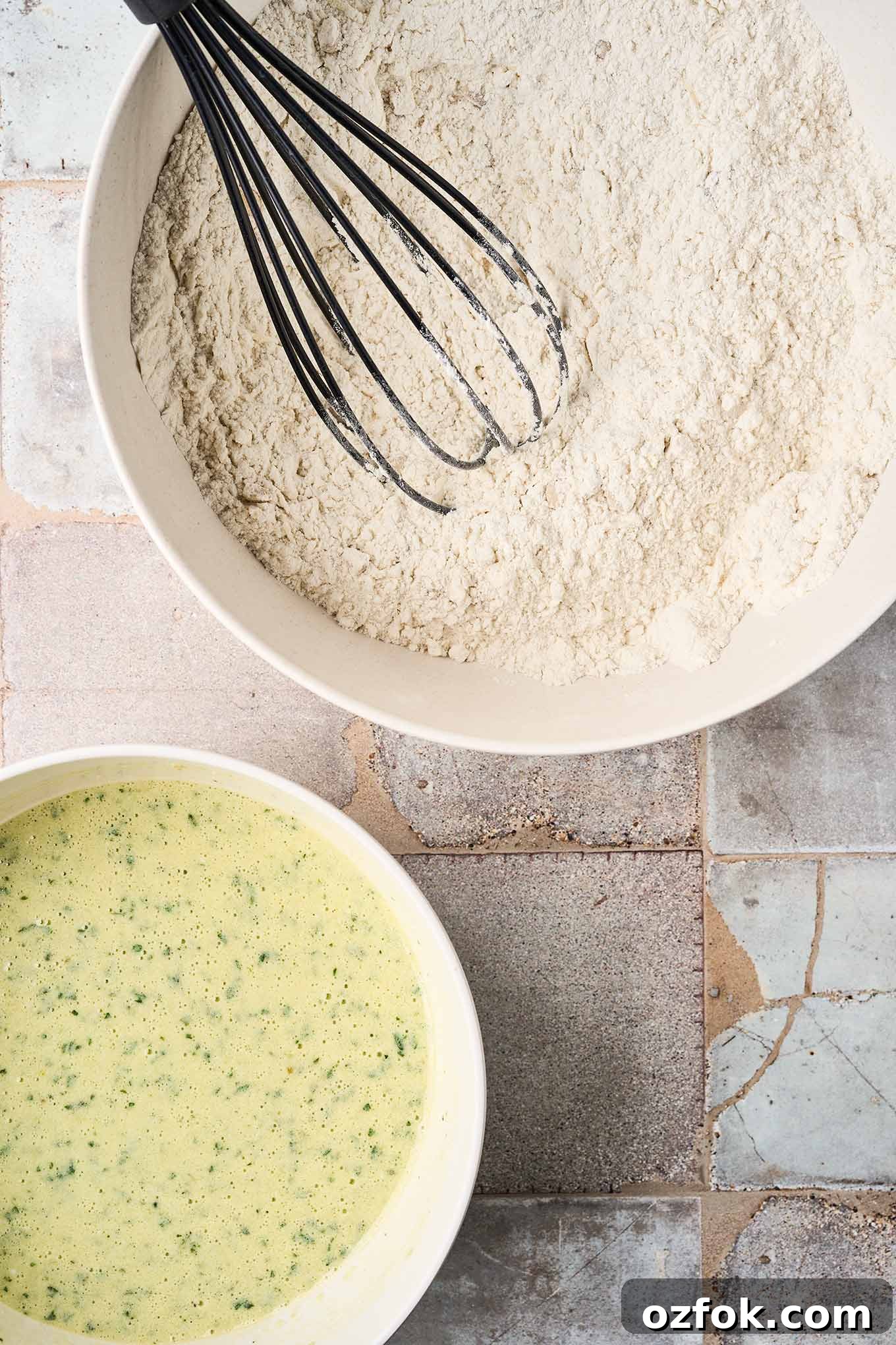 The dry ingredients for savory zucchini bread in a mixing bowl with a whisk beside a smaller mixing bowl containing the wet ingredients, ready to be combined.