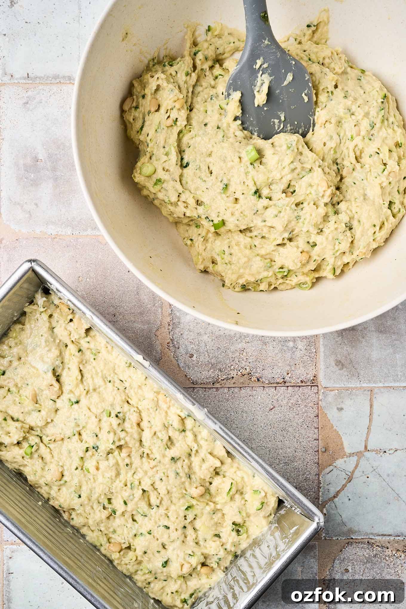 Pesto zucchini bread batter in a bowl with a spatula beside a loaf pan already filled and ready for baking.