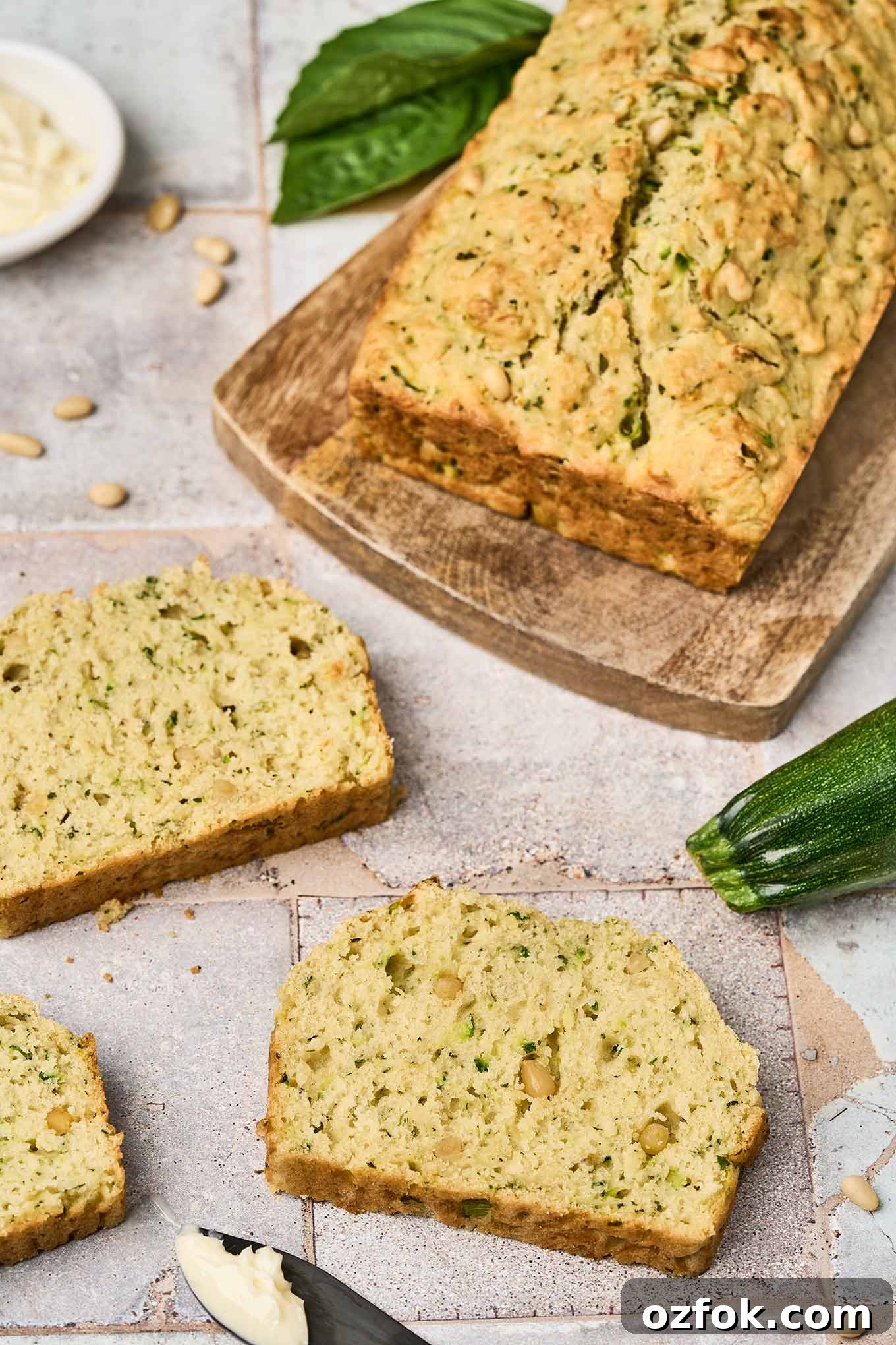 A close view of slices of savory zucchini bread on a tile countertop, a knife with butter on it, and a cutting board with a loaf of zucchini bread on it.