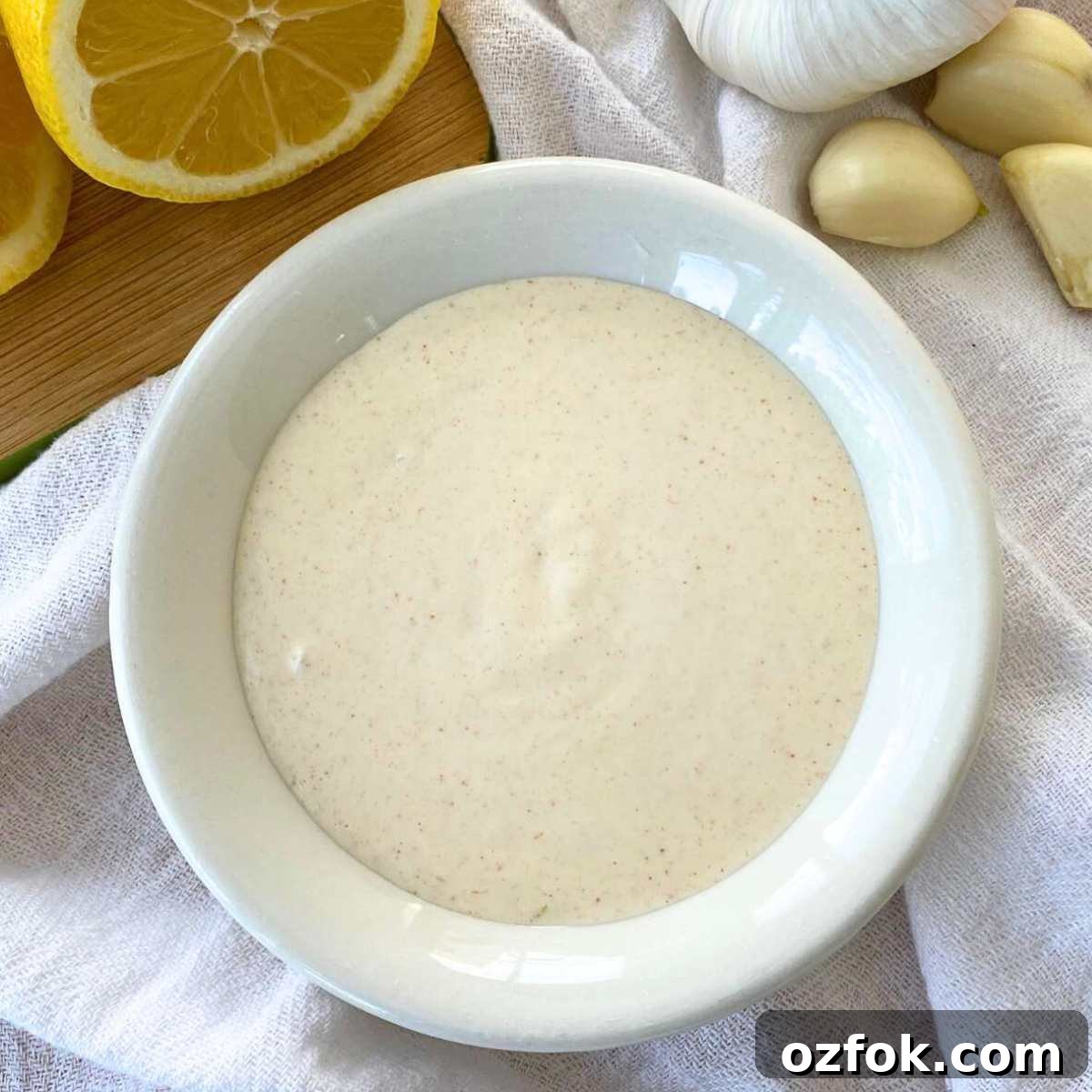 Lemon Garlic Aioli: Homemade Creamy Sauce Recipe 2 Lemon garlic aioli sauce in a bowl with a cut lemon and garlic cloves behind it.