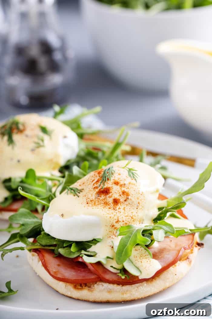 Classic Eggs Benedict Recipe: How to Make Perfect Hollandaise and Poached Eggs 2 Eggs benedict with canadian bacon and arugula on a white plate.