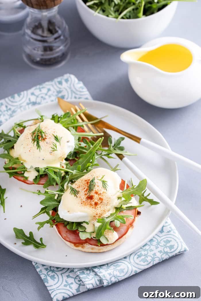 Classic Eggs Benedict Recipe: How to Make Perfect Hollandaise and Poached Eggs 11 White plate with eggs benedict set next to a container of hollandaise.