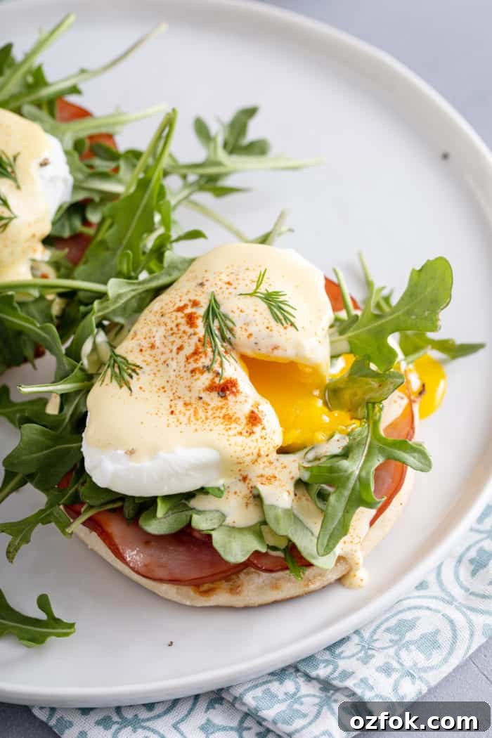 Classic Eggs Benedict Recipe: How to Make Perfect Hollandaise and Poached Eggs 3 Plated eggs benedict with the egg cut so the yolk is running out over the arugula, candian bacon, and english muffin.