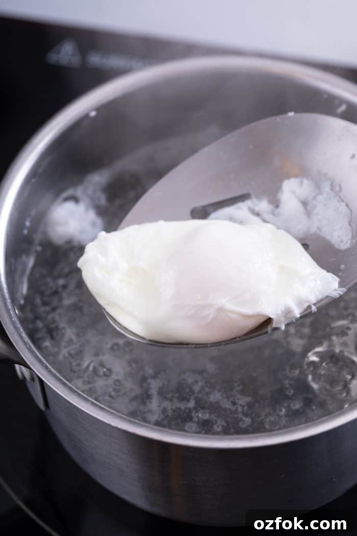 Classic Eggs Benedict Recipe: How to Make Perfect Hollandaise and Poached Eggs 7 Freshly poached egg being removed from simmering water with a slotted spoon.