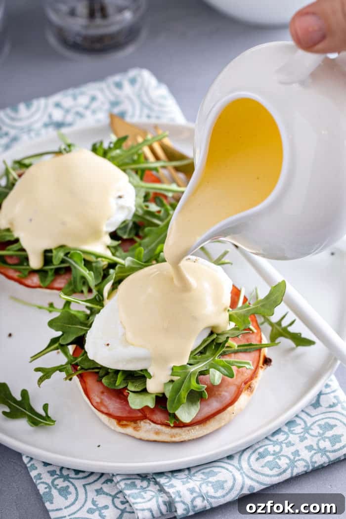 Classic Eggs Benedict Recipe: How to Make Perfect Hollandaise and Poached Eggs 9 Hollandaise sauce being poured over eggs benedict on a white plate.