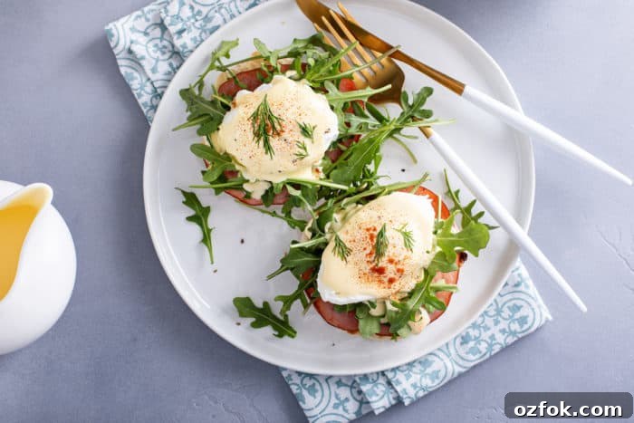 Classic Eggs Benedict Recipe: How to Make Perfect Hollandaise and Poached Eggs 10 Overhead view of eggs benedict next to a fork and knife on a white plate, set on a blue napkin.