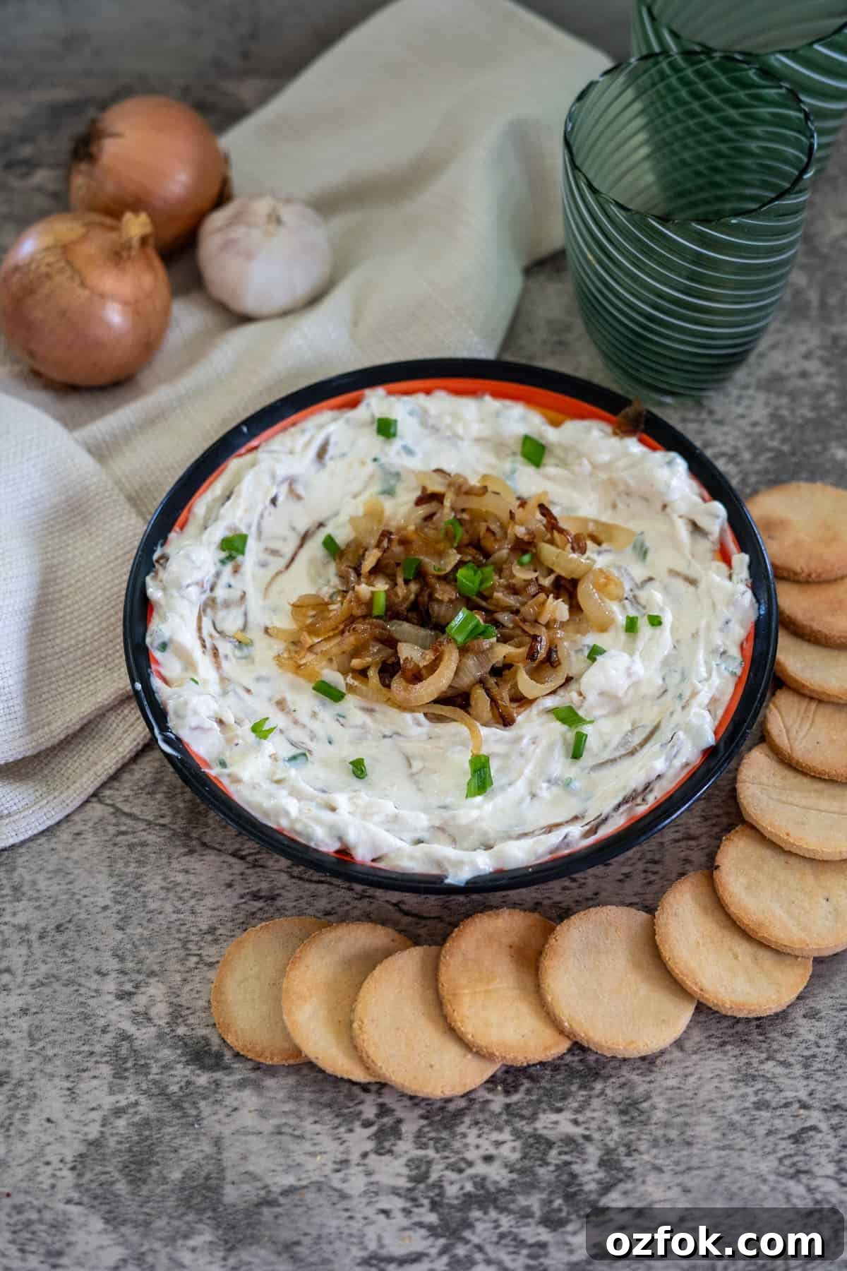 Keto French Onion Dip Recipe for Low-Carb Snacking 2 A bowl of onion dip garnished with caramelized onions and chives, surrounded by round crackers. Onions, garlic, and a cloth napkin are in the background alongside two green glasses.