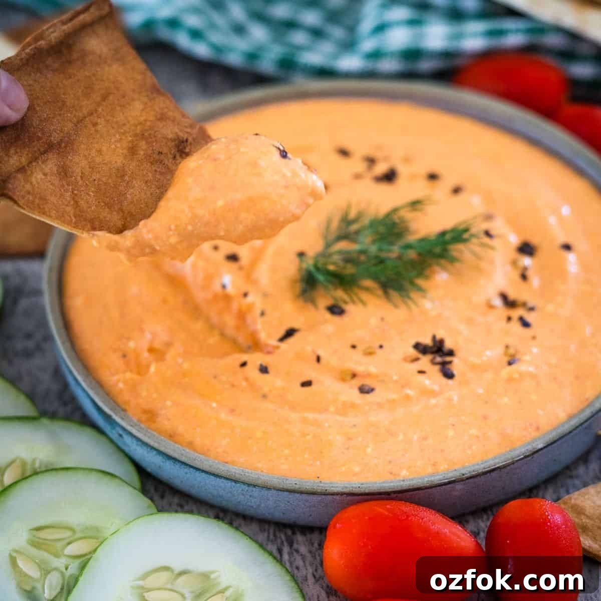 Keto French Onion Dip Recipe for Low-Carb Snacking 11 A hand dipping a piece of pita into a bowl of creamy orange dip garnished with a sprig of dill. Sliced cucumbers and cherry tomatoes are visible around the bowl.