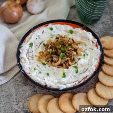 Keto French Onion Dip Recipe for Low-Carb Snacking 14 A bowl of onion dip garnished with caramelized onions and chives, surrounded by round crackers. Onions, garlic, and a cloth napkin are in the background alongside two green glasses.