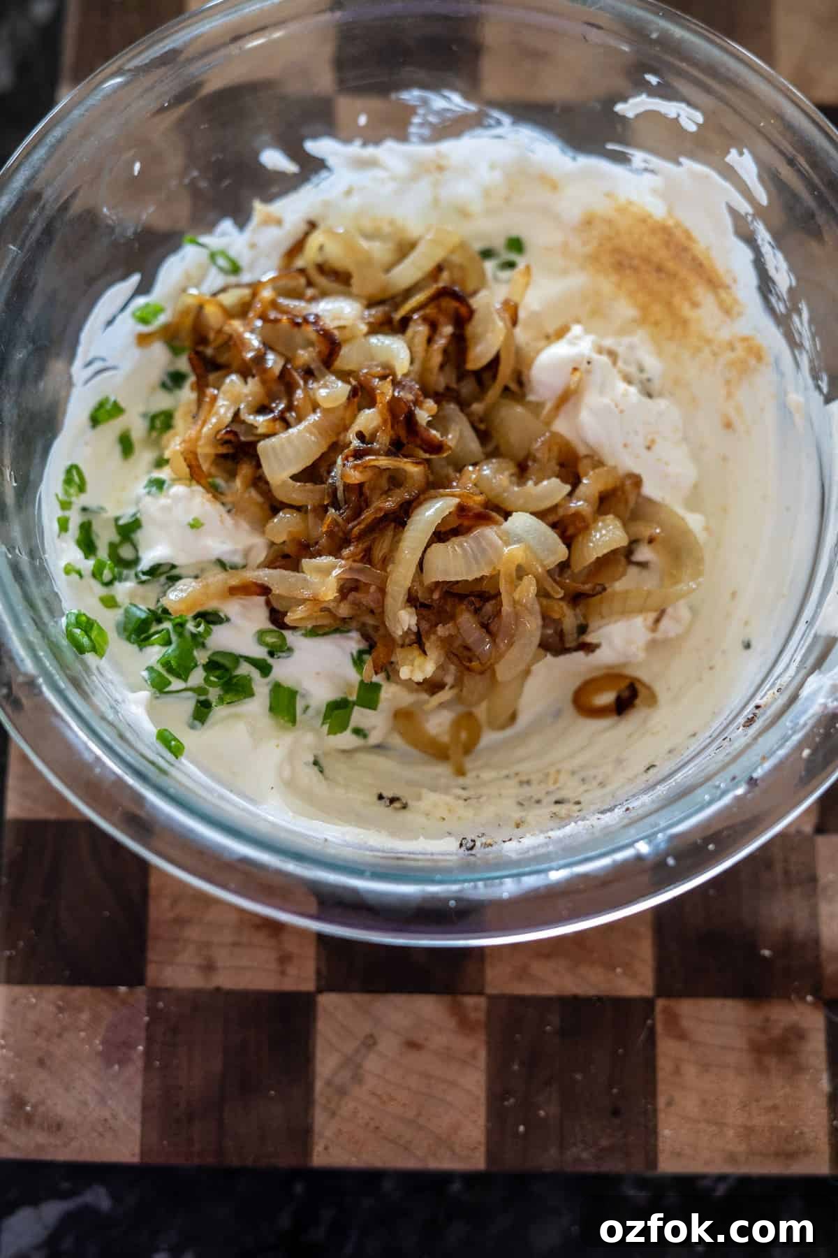 Keto French Onion Dip Recipe for Low-Carb Snacking 6 Glass bowl with sour cream, caramelized onions, chopped chives, and seasoning on a wooden cutting board.