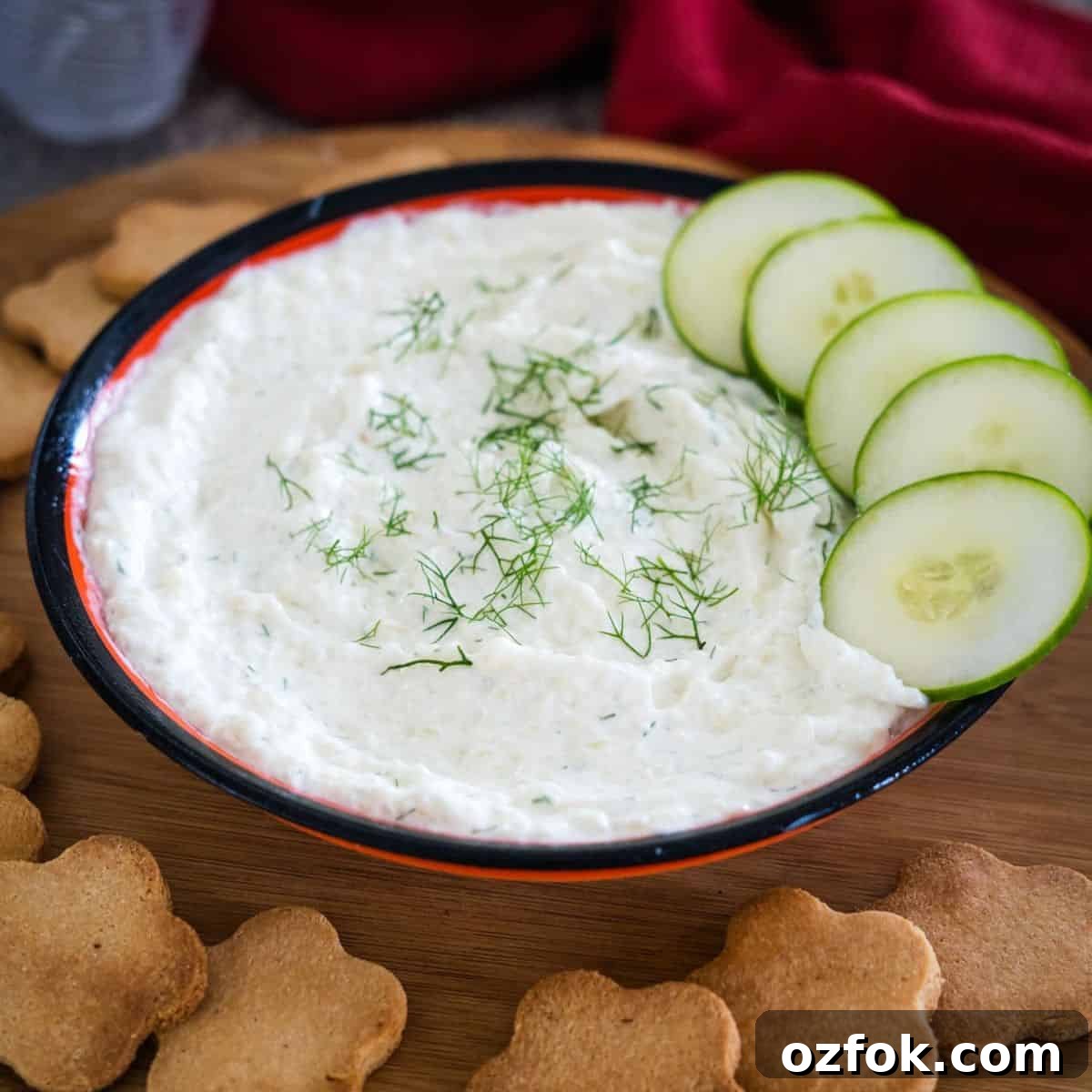 Keto French Onion Dip Recipe for Low-Carb Snacking 10 A bowl of creamy dip garnished with dill, accompanied by cucumber slices and flower-shaped crackers on a wooden board.
