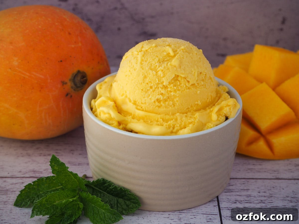 Creamy Mango Ice Cream Recipe: Fresh Mango Frozen Dessert 2 Mango ice cream in a small bowl, with fresh mint on the side and fresh mangoes in the background.