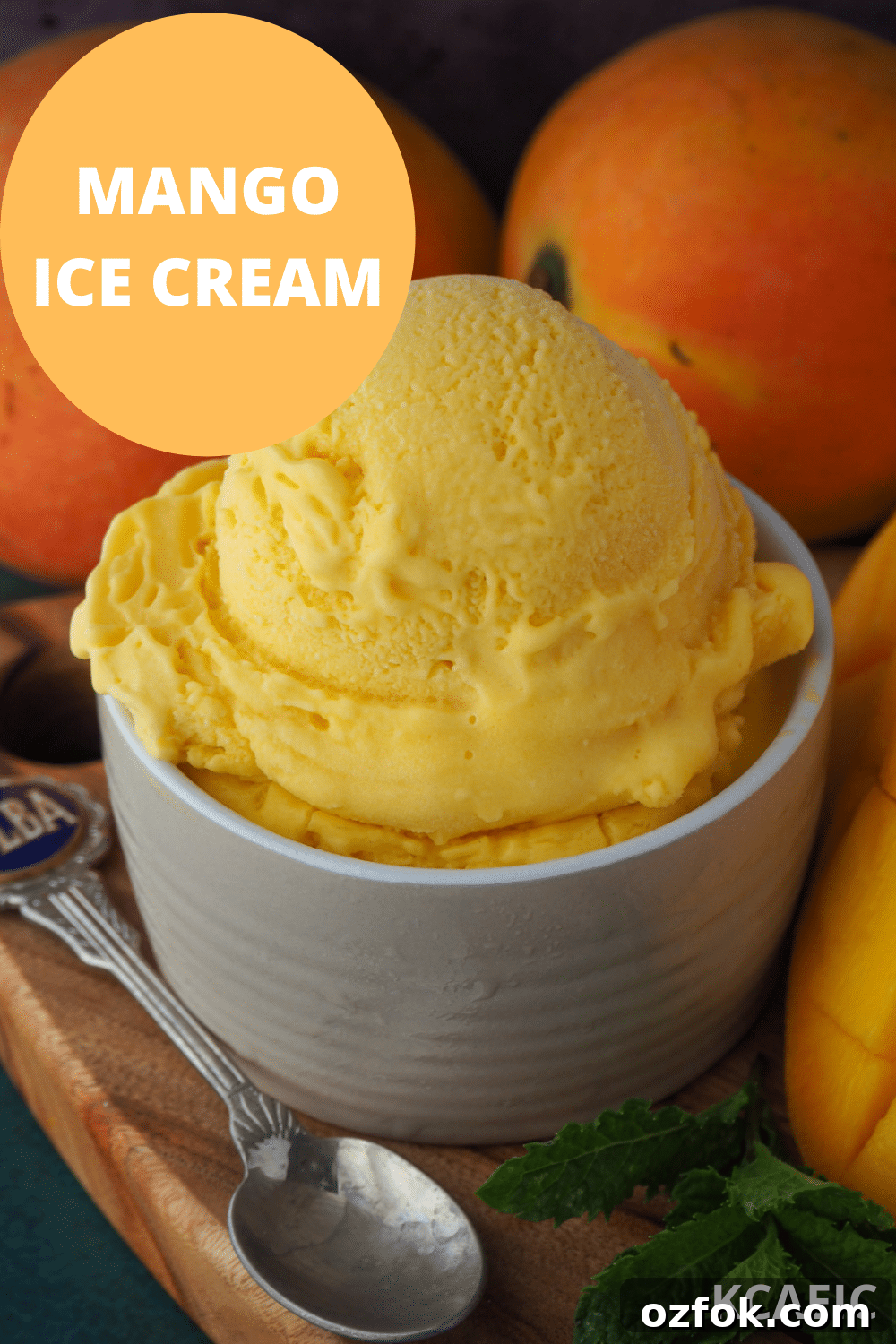 Creamy Mango Ice Cream Recipe: Fresh Mango Frozen Dessert 17 Mango ice cream in a small bowl, on a chopping board with a vintage spoon and mint in the foreground, and fresh mangos in the background.