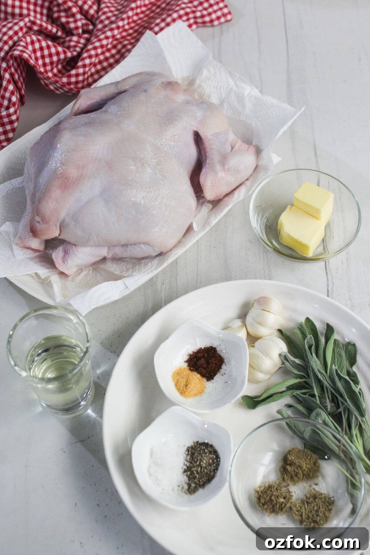 Crispy Air Fryer Spatchcock Chicken Recipe for Juicy Results 3 Uncooked whole chicken on a plate with butter, garlic, fresh sage, spices, herbs, and a glass of liquid on a white table.