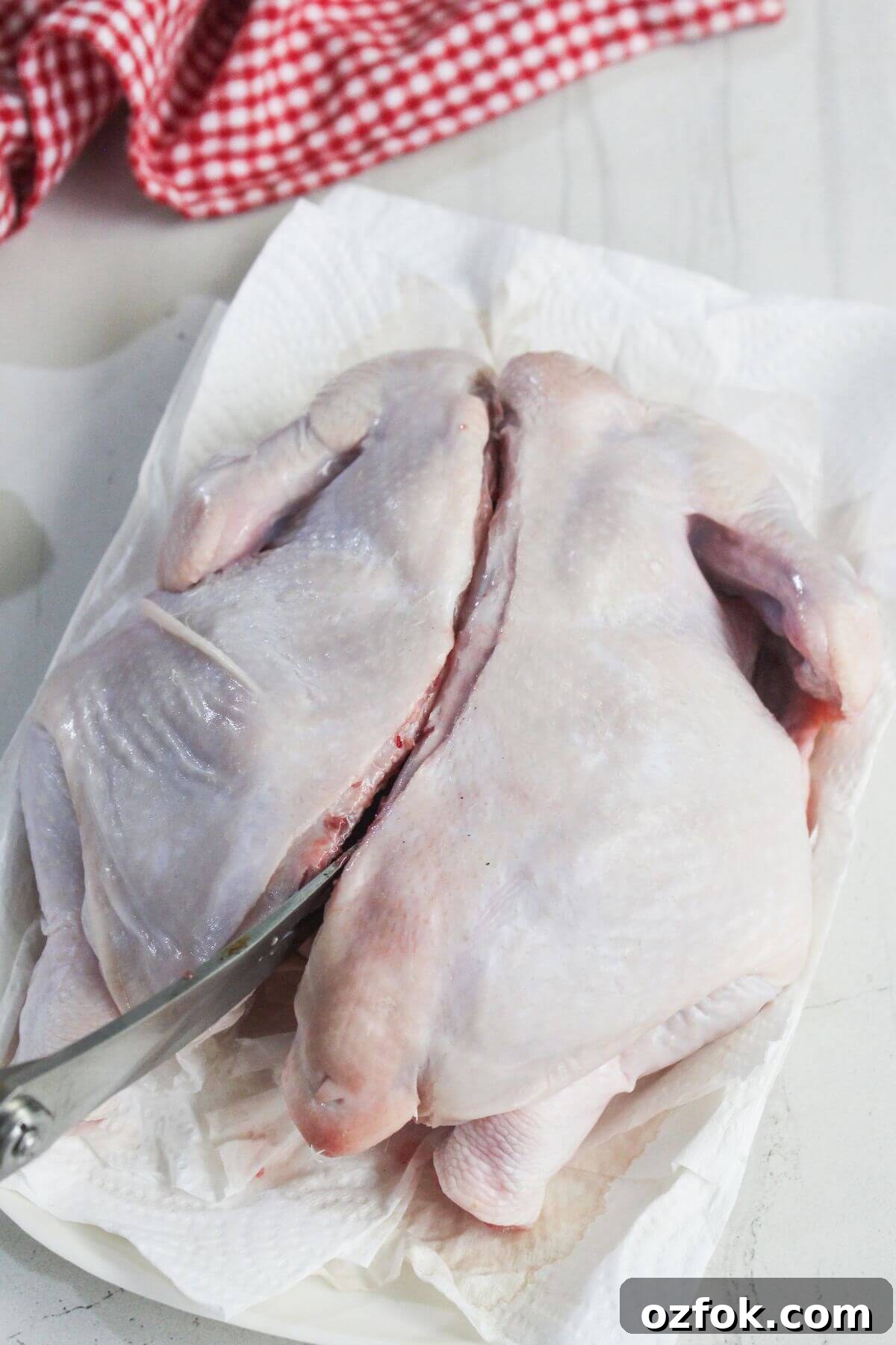 Crispy Air Fryer Spatchcock Chicken Recipe for Juicy Results 4 A raw whole chicken is being butterflied with a knife on a paper towel-lined surface, next to a red and white checkered cloth.