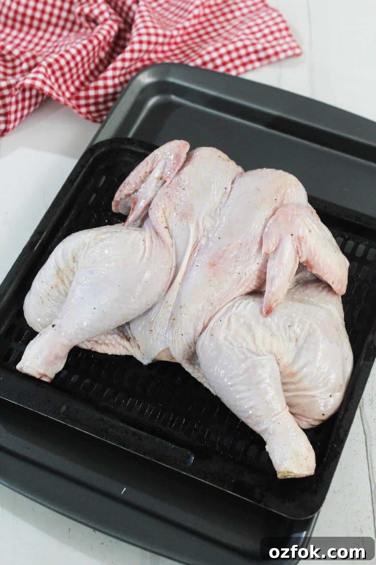 Crispy Air Fryer Spatchcock Chicken Recipe for Juicy Results 7 Raw spatchcocked chicken placed on a black roasting tray, with a red and white checkered cloth in the background.