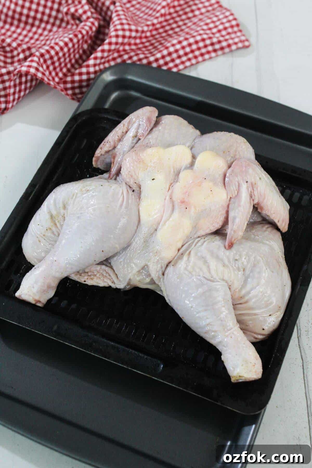 Crispy Air Fryer Spatchcock Chicken Recipe for Juicy Results 8 A raw spatchcocked chicken is placed on a black roasting tray, with a red and white checkered cloth in the background.