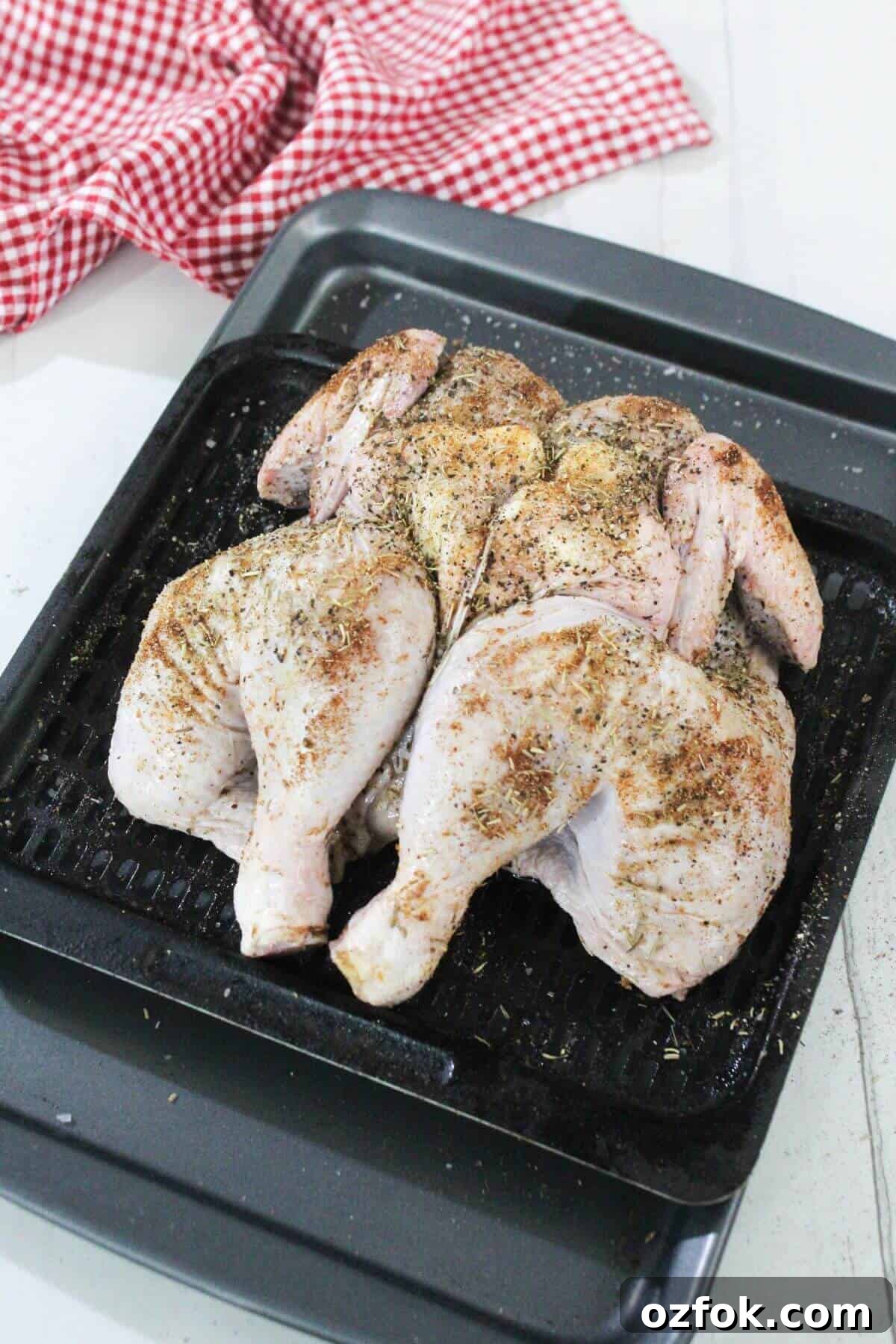 Crispy Air Fryer Spatchcock Chicken Recipe for Juicy Results 9 A raw, spatchcocked chicken seasoned with spices sits on a black roasting pan, with a red and white checkered cloth in the background.