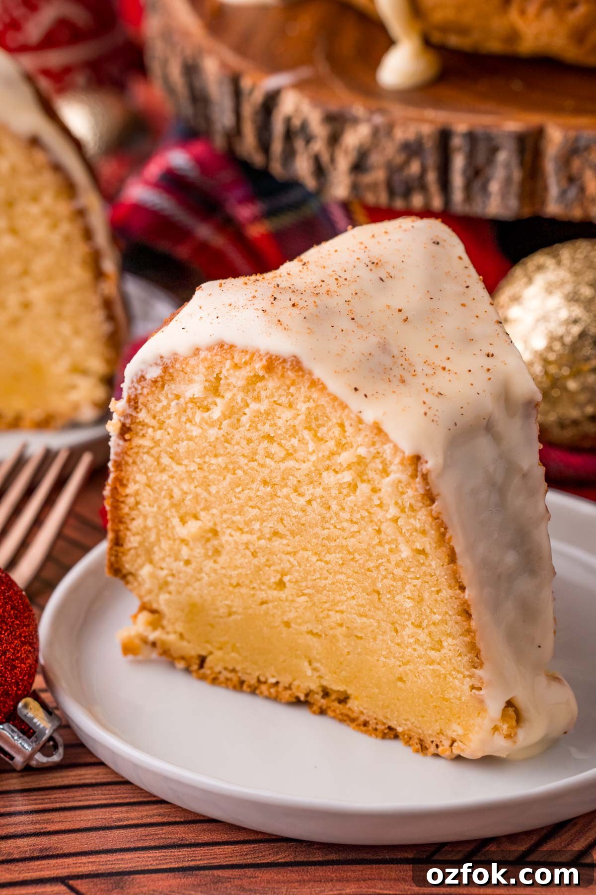 Close up photo of a slice of eggnog pound cake on a white plate with the rest of the cake in the background decorated for the holidays.
