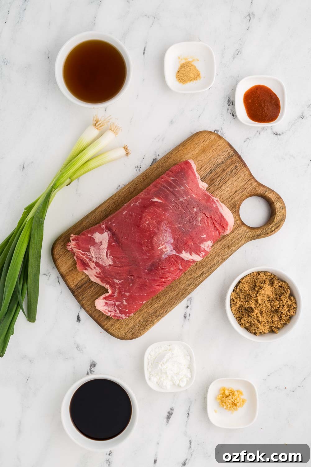 Ingredients for shredded Mongolian beef recipe on a counter: 1 1/2 pounds of flank steak, 1/4 cup soy sauce, 1/4 cup beef broth, 1/4 cup dark brown sugar, 1 teaspoon jarred minced garlic, 1/4 teaspoon powdered ginger, 1 teaspoon Sriracha sauce, 2 tablespoons corn starch, and 1/2 bunch green onions