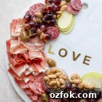 A large marble stone with the word "love" surrounded by deli meats, grapes, olives, nuts, and slices of green apple.