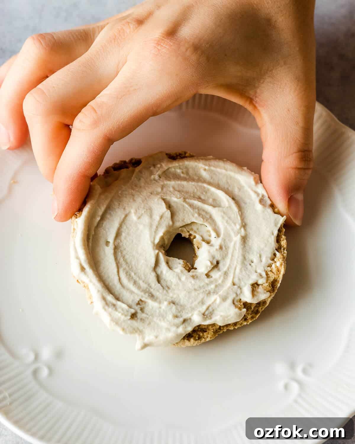 Homemade Vegan Cream Cheese Recipe — Smooth & Tangy Plant-Based Spread 3 A hand is grabbing half a bagel with cream cheese on a white plate.